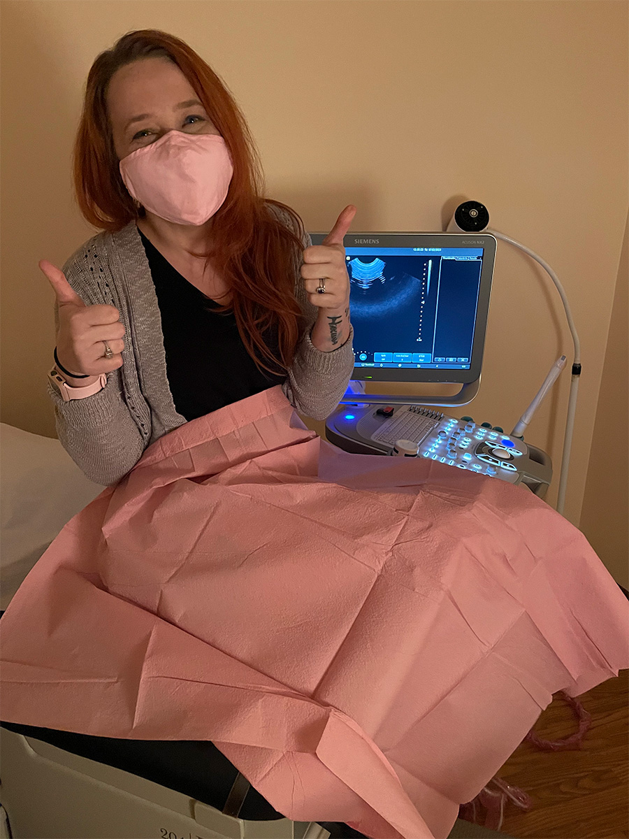 Woman sitting on exam room table giving a thumbs up while smiling and wearing a mask.