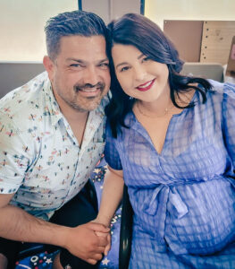 A happy pregnant couple sitting down, smiling, and holding hands.