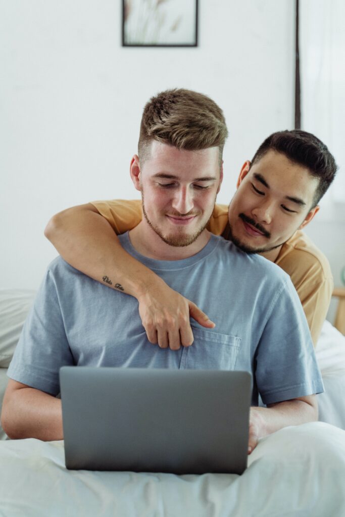 Same-sex interracial male couple smiling, sitting in bed, and looking at laptop.