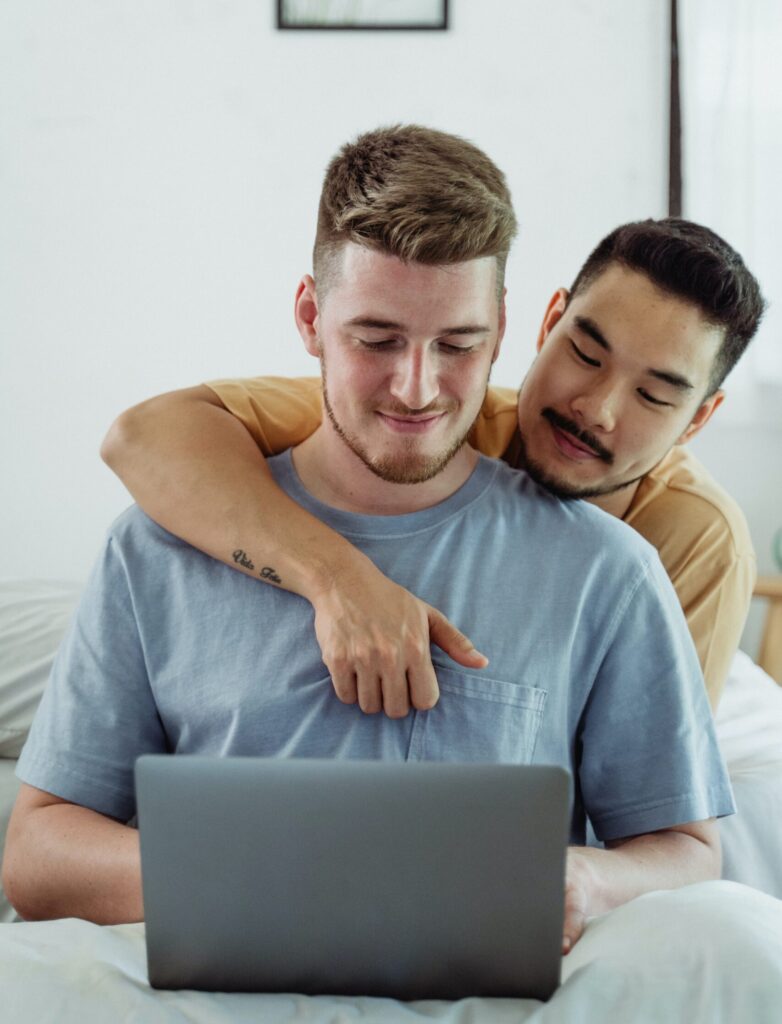 Same-sex interracial male couple smiling, sitting in bed, and looking at laptop.