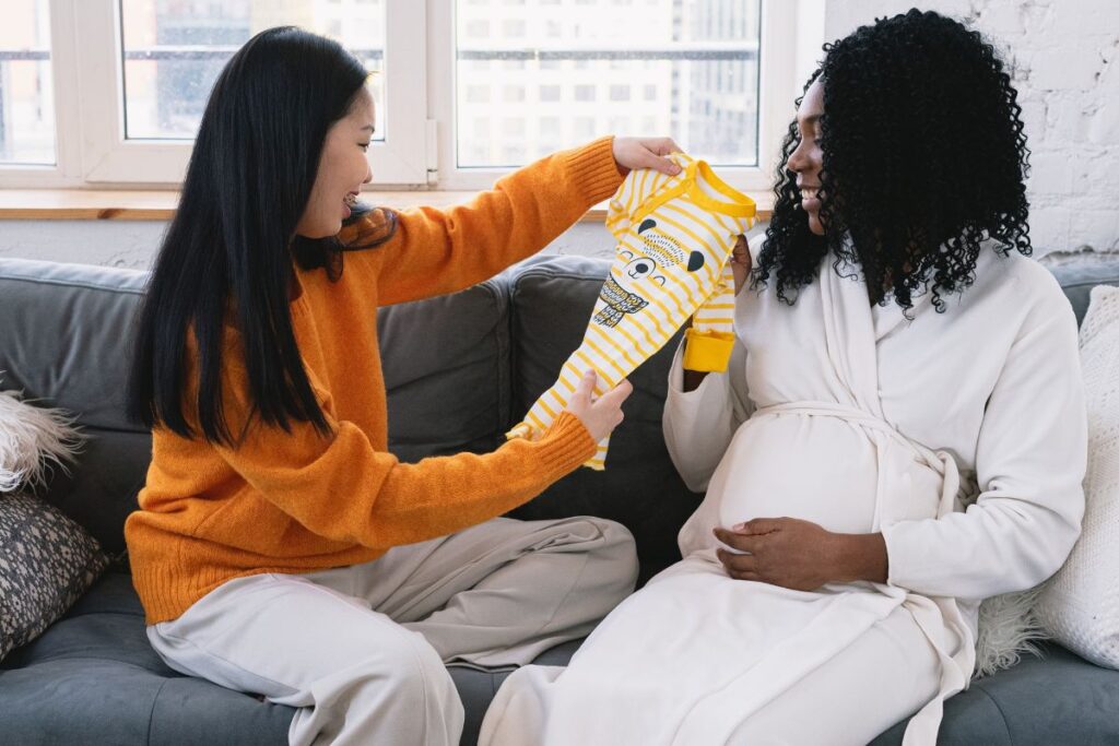 a pregnant female same-sex couple sitting on couch looking a baby clothes
